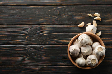 Wooden bowl of fresh garlic bulbs, slices on wooden background, top view. Space for text