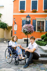 Couple in love in old city center. Pretty young girl with disease on a wheelchair and her lovely man, sitting on the stairs. Stylish orange ancient building on the background