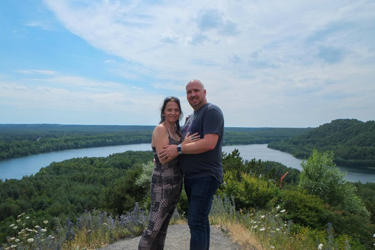 Loving Couple Of 30-40 Years Old Outdoors Hugging And Standing Tall On A Hill With A River Running Through The Woods The Background