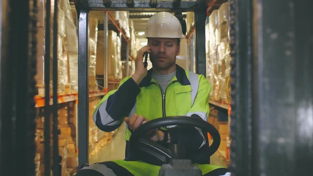 Portrait of male worker talking on mobile phone while driving forklift in warehouse.