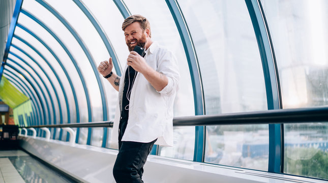 Positive male meloman happy from received voice message with good news listening during free time for music hobby, young man in casual outfit and headphones dancing indoors during radio entertainment