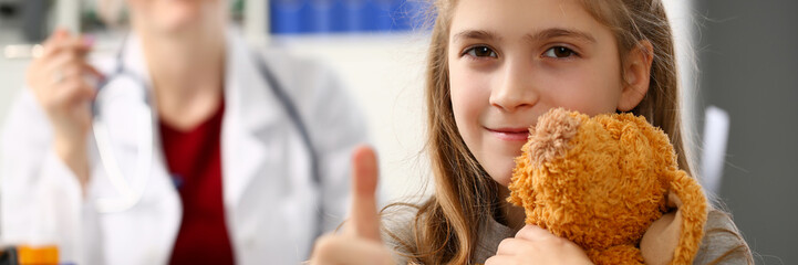 Happy little child girl hold teddy bear and thumbs up in hospital female excel doctor office.