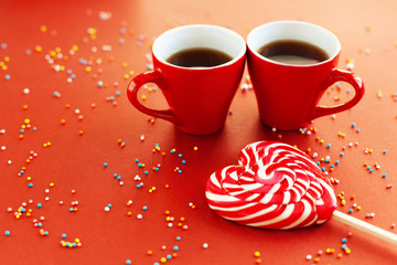 Two cups of hot drinks and lollipop in the shape of heart on bright red background. Romantic...