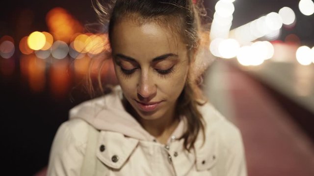 slow motion Closeup of beautiful young woman using smartphone walking outdoors night city, wind blowing hair. female model messaging, texting, swiping screen on mobile phone on night city lights