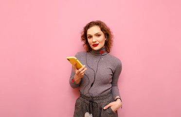 Attractive girl in casual clothes, bright makeup and curly red hair isolated on a pink pastel background with a smartphone in her hand, looks into the camera and listens to music in headphones.