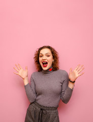 Studio portrait of joyful lady in casual clothes and with curly red hair on pastel pink background, looks into camera with surprised face and rejoices. Cheerful girl emotionally rejoices. Vertical