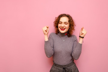 Positive teen girl with curly red hair and in a gray sweater stands with her eyes closed on a pink...