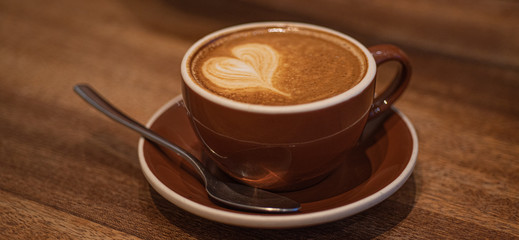Cup of cappuccino coffee with heart art foam and spoon on wooden table on blurred background. Wide photo.