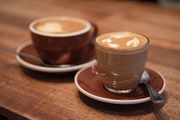 Glass Cup of cortado coffee next to blurred cup of cappuccino on wooden table. Close up view.
