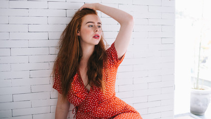 Confident woman with bright hair in red dress sitting near white brick wall