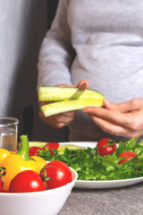 Unrecognizable pregnant woman cutting cucumber for salad in the kitchen. Cucumber, cherry tomatoes and fresh salad are already on the white plate. Healthy food and lifestyle concept.