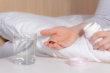 Unrecognizable woman sitting on bed and holding sleeping pills for insomnia before intake. Glass of water is on bedside table. Jet lag effect. Snow-white bedding.