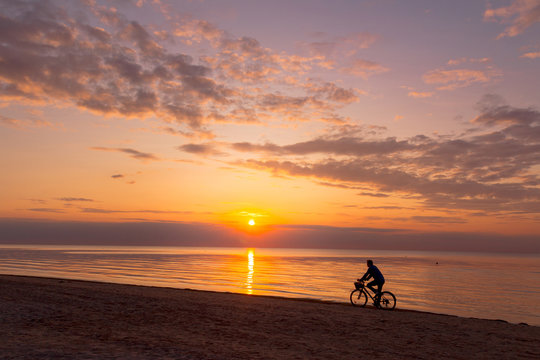 Beautiful sunset sky with clouds over Baltic sea beach coastline. Jurmala. Latvia. Small silhouette of man on a bicycle cycling along a coast. - Powered by Adobe