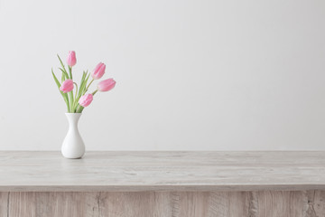 pink tulips in white ceramic vase on wooden table on background white wall