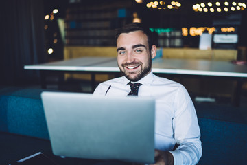 Portrait of cheerful male entrepreneur sitting in corporate workspace and smiling at camera during time for bitcoin analyzing of crypto currency exchange on trade websites, laptop investment