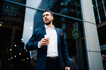 Tired businessman in classical suit with coffee to go and smartphone looking away walking in street in downtown