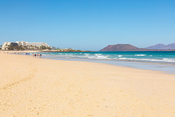 Fototapeta na wymiar Beach hotel on Fuerteventura canary islands with the beach in front and the sea with blue azure aquamarine turquoise colors and a vulcano in the background blue sky
