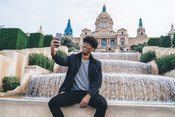 Cheerful black man taking selfie at fountain edge with cellphone