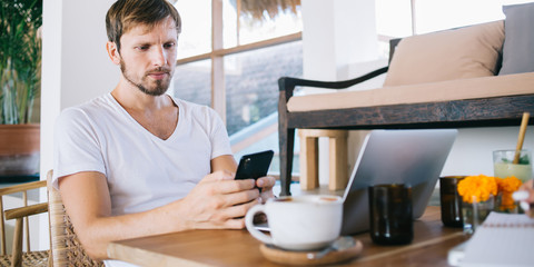 Serious male using laptop and texting on mobile and drinking brew