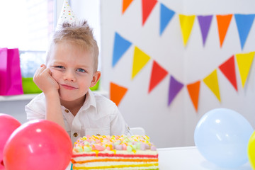 Fototapeta na wymiar Unhappy blonde caucasian boy with boring face near birthday rainbow cake. Festive colorful background. Bad birthday party.