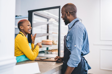 Black seller in uniform smiling and speaking to colleague about cake