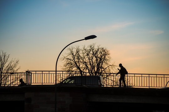   Silhouette of young boy walking on bridge under the river with smartphone in hands by sunset