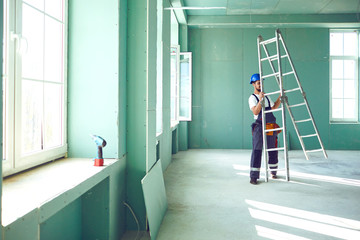 A builder standing on a ladder installs drywall at a construction site