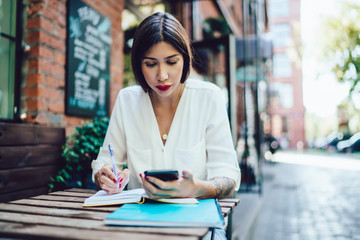 Attractive female blogger reading received email on cellular connected to 4g wireless for browsing internet during learning in street cafeteria, hipster girl chatting researching information for test