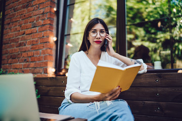 Portrait of female freelancer sitting in cafe terrace with modern netbook for making distance job and calling via cellular application, Latino woman holding memo textbook for making time management