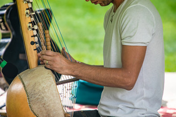 Naklejka premium Midsection of male artist in white t-shirt performing traditional wooden harp kora while sitting during event in city against lawn
