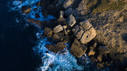 Top view of the coast. In the image we can see the blue sea with some rocks on a colorful sunset.