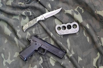 Handgun lies with brass knuckles and knife on camouflage military uniform close up. Concept of...