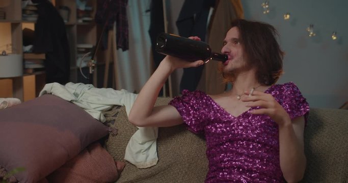 Drunky man transgender in fashionable dress sitting on sofa at home and smoking cigarette. Man transgender coming back from party, drinking wine from bottle and singing. Room background .