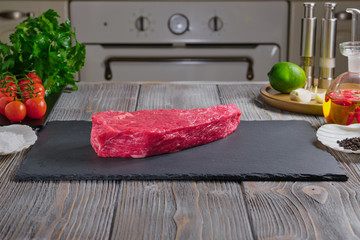 Marbled Beef Steak a rectangular plate of black color ardesia material, on a wooden table. Spices, parsley, lime on a wooden table. Red hot peppers, cherry tomatoes, garlic.