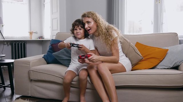 Young woman and 5 age boy are sitting on a couch in light living room and playing online video games on wireless controllers at home