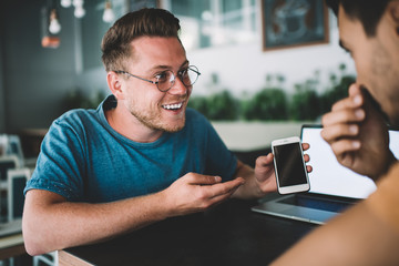 Excited young male businessman sharing good news with colleague on smartphone in cafe