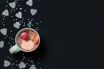 Heart shaped confetti with a rosebud in a white cup on black background. Top view, copy space.