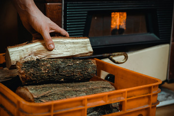 Focus on foreground with man's hand grabing a piece of firewood from basket, flame in stove in blurred background.