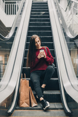 Young pretty woman blogger with smartphone, cup of coffee and shopping bags in city mall
