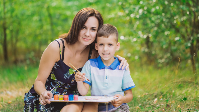 Portrait of happy family of european mother and little son having fun outdoors in summer green park or backyard. Little boy learnig to paint pictures. Horizontal color photography.