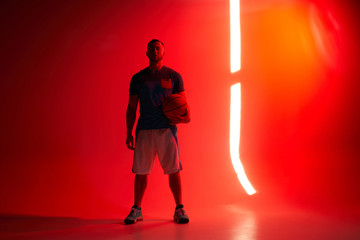 Young athletic man, basketball player stands holding ball in one hand with back lights and red background