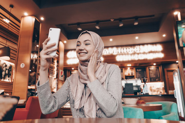 smiling muslim woman looking at camera phone in cafe