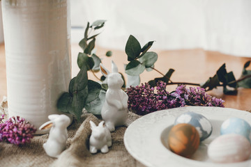 Stylish Easter eggs on vintage plate, white bunny rabbits and lilac flowers on rustic fabric on wooden table. Rural easter composition of natural dyed eggs and spring purple flowers