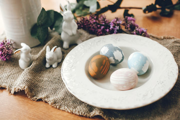 Stylish Easter eggs on vintage plate, white bunny rabbits and lilac flowers on rustic fabric on wooden table. Rural easter composition of natural dyed eggs and spring purple flowers
