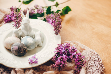 Stylish Easter eggs on vintage plate, white bunnies and lilac flowers on fabric on wooden table. Rural  natural dyed easter eggs and spring flowers. Space for text. Holiday decor