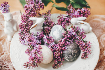 Happy Easter. Stylish Easter eggs on vintage plate, white bunnies and lilac flowers on fabric on wooden table. Rural composition of colorful natural dyed easter eggs and spring flowers