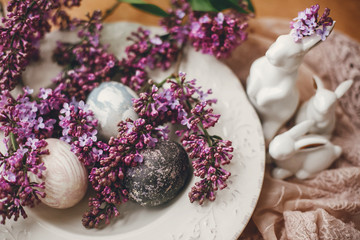 Happy Easter. Stylish Easter eggs on vintage plate, white bunnies and lilac flowers on fabric on wooden table. Rural composition of colorful natural dyed easter eggs and spring flowers