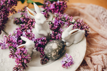 Stylish Easter white bunnies, modern easter egg on vintage plate,  and lilac flowers on rural fabric. Natural dyed easter egg and spring flowers. Space for text. Holiday decor