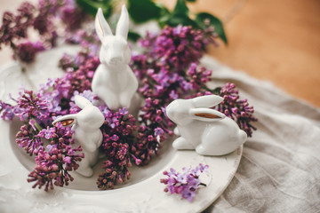 Stylish Easter white bunnies on vintage plate and lilac flowers on rural fabric. Ceramic rabbits decorations and spring flowers. Space for text. Holiday decor