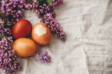 Fototapeta na wymiar Happy Easter. Modern colorful Easter eggs on rustic linen fabric and purple lilac flowers. Rural still life. Space for text. Natural dyed easter eggs. Stylish holiday table decor